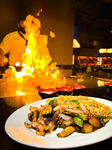 A plate of grilled veggies and rice in the foreground with a chef standing behind a wall of bright flames in the background.