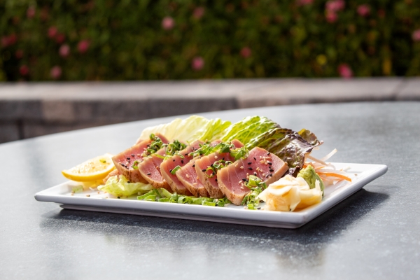 A white, rectangular plate on a gray outdoor table with slices of pink ahi tuna garnished with ginger, wasabi, lettuce leaves, and lemon wedges. 