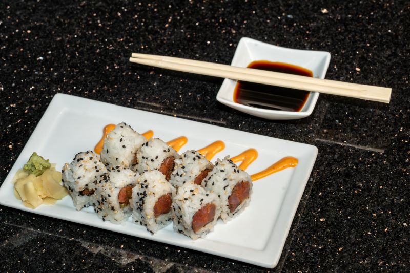 A plate of sushi with ginger, wasabi, and a drizzle of spicy mayo on a white plate. The sushi contains rice and seeds on the outside and tuna on the inside, and there's a small dish of soy sauce on the side with chopsticks.  Let the servers know you’re a sushi beginner and they can give you the best recommendations for what rolls to start with. 