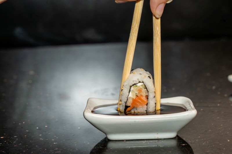 A close up image of chopsticks squeezing and dipping a piece of sushi into a small white dish filled with soy sauce. A great tip for sushi beginners is to dip into different sauces to see which combinations you like best. 