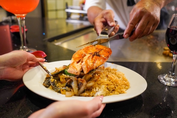 A close-up of a chef’s hands using a spatula to place a seared piece of salmon on a customer’s plate of grilled veggies and rice. 