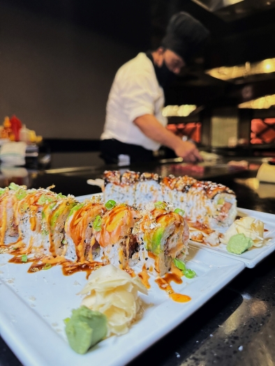 Two colorful sushi rolls on white plates in the foreground with a chef in a white chef’s jacket out of focus standing at a grill top in the background. Bring Mom to Shogun this Mother's Day.