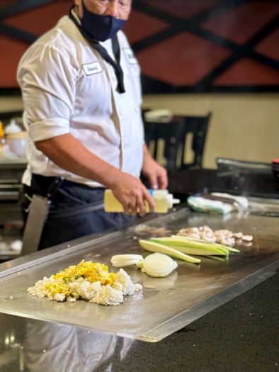 A chef in a white chef coat and black mask stands behind a grill top, holding a bottle of oil over a pile of rice, fresh onions, zucchini, and mushrooms. 