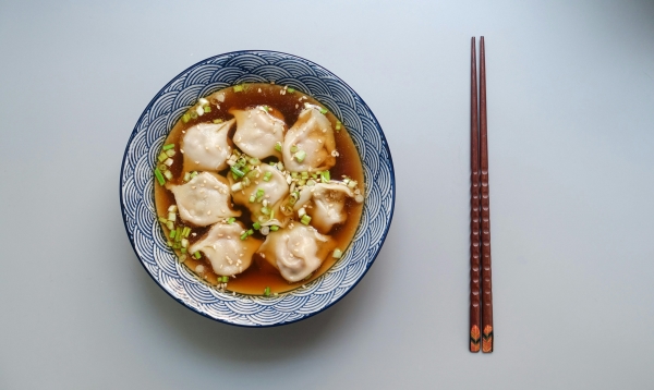 A blue and white patterned bowl with wonton soup and scallion garnishes on a gray background next to a pair of dark brown chopsticks. 