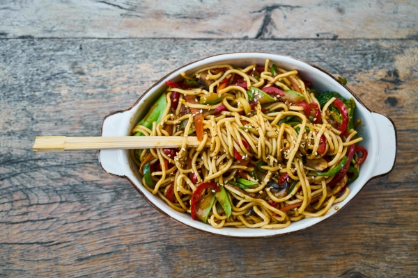 An overhead shot of an oblong dish with Chinese noodles and vegetables against a wooden background. Noodles are wrapped around a pair of chopsticks that are laid on top of the plate. 