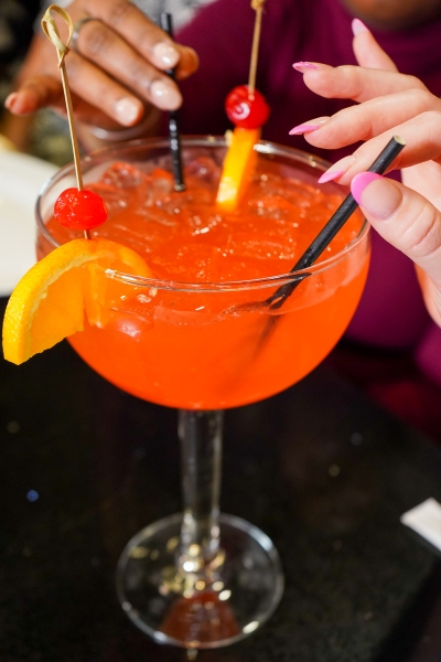 A close-up of a bright orange cocktail in a large glass garnished with orange slices and cherries with two women holding black straws. 