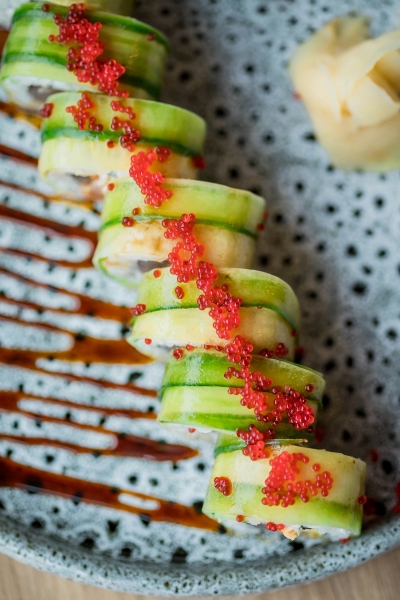 A shot from above of staggered sushi rolls with an outer layer of avocado topped with bright red roe on a gray and black spotted dish with a drizzle of eel sauce. 