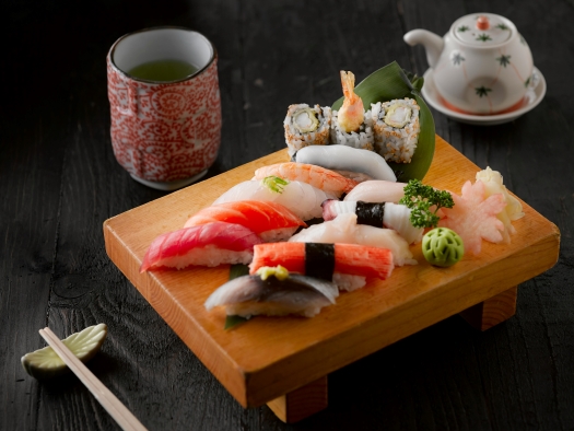 A wooden stand with an array of nigiri and sashimi sushi, wasabi, ginger, and three sushi rolls beside a cup of tea, a teapot, and a pair of chopsticks on a chopstick rest.  