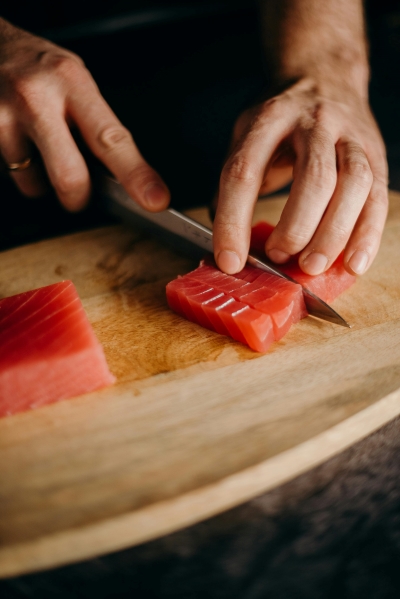 Hands use a knife to slice into a deep pink filet of raw tuna fish against a wooden cutting board.  