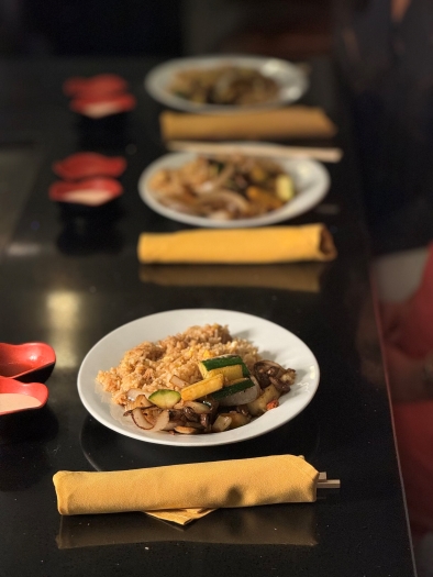 Three plates of rice and veggies beside silverware wrapped in yellow linen napkins on a long black table. 