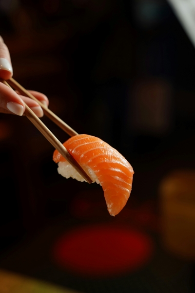 A hand holding piece of salmon sashimi with a pair of chopsticks against an out of focus dark background.  