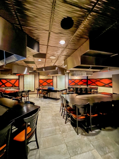 An empty restaurant with large teppanyaki tables surrounded by chairs with orange cushions. 