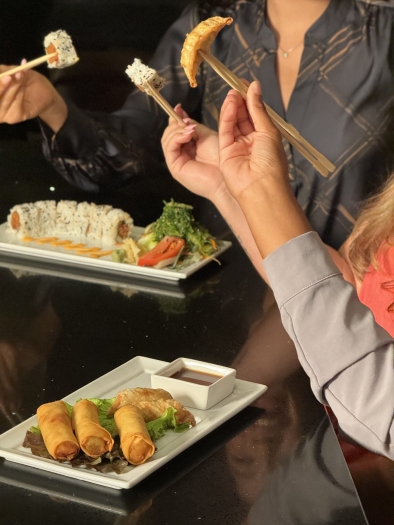 Three women holding chopsticks at a table with a plate of sushi and a plate of fried spring rolls. Practice your Japanese culinary etiquette at Shogun Japanese Steakhouse and Sakura Sushi.  