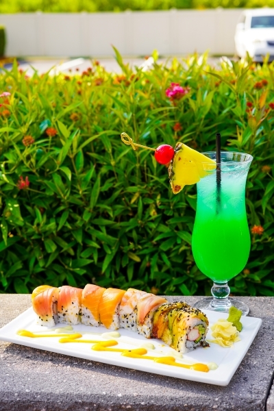 On an outdoor stone slab, a white rectangular plate with colorful sushi rolls sits beside a bright green cocktail in a hurricane glass with a pineapple garnish.  