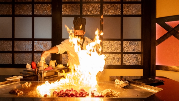 A chef in a tall black hat with a black mask stands behind a grill table with a tall flame. 