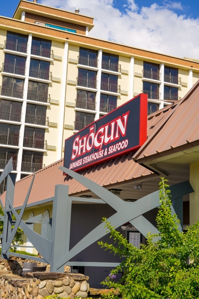 The exterior a building with a brown roof and decorative archways resembling samurai swords beneath a large sign reading ” Shogun Japanese Steakhouse & Seafood.” A hotel rises up in the background. You can experience the health benefits of Japanese cuisine at Shogun.