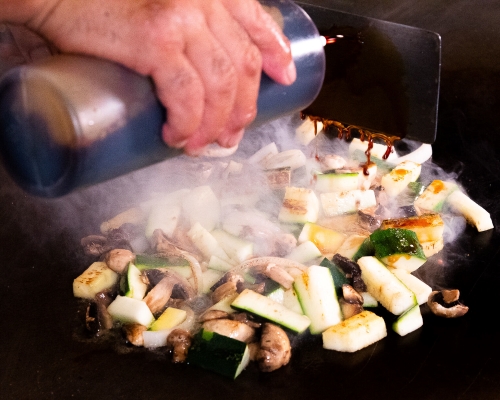 A close-up of a hand squeezing brown sauce onto a steaming pile of mushrooms and zucchini on a grill top. 