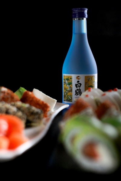 Out of focus sushi in the foreground with an in-focus blue bottle of sake in the background against a black backdrop.  