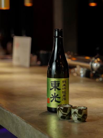 A dark bottle of sake with a green shiny label sits beside two sake cups on a cement bar counter.