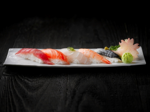 A long, rectangular white plate with five pieces of colorful sashimi plated with a small ball of green wasabi and a flower cut out of pickled ginger. 