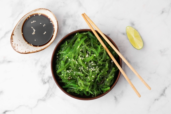 An overhead shot of a bowl with bright green seaweed salad, chopsticks, a small dish of soy sauce, and a lime wedge against a white marble backdrop. 